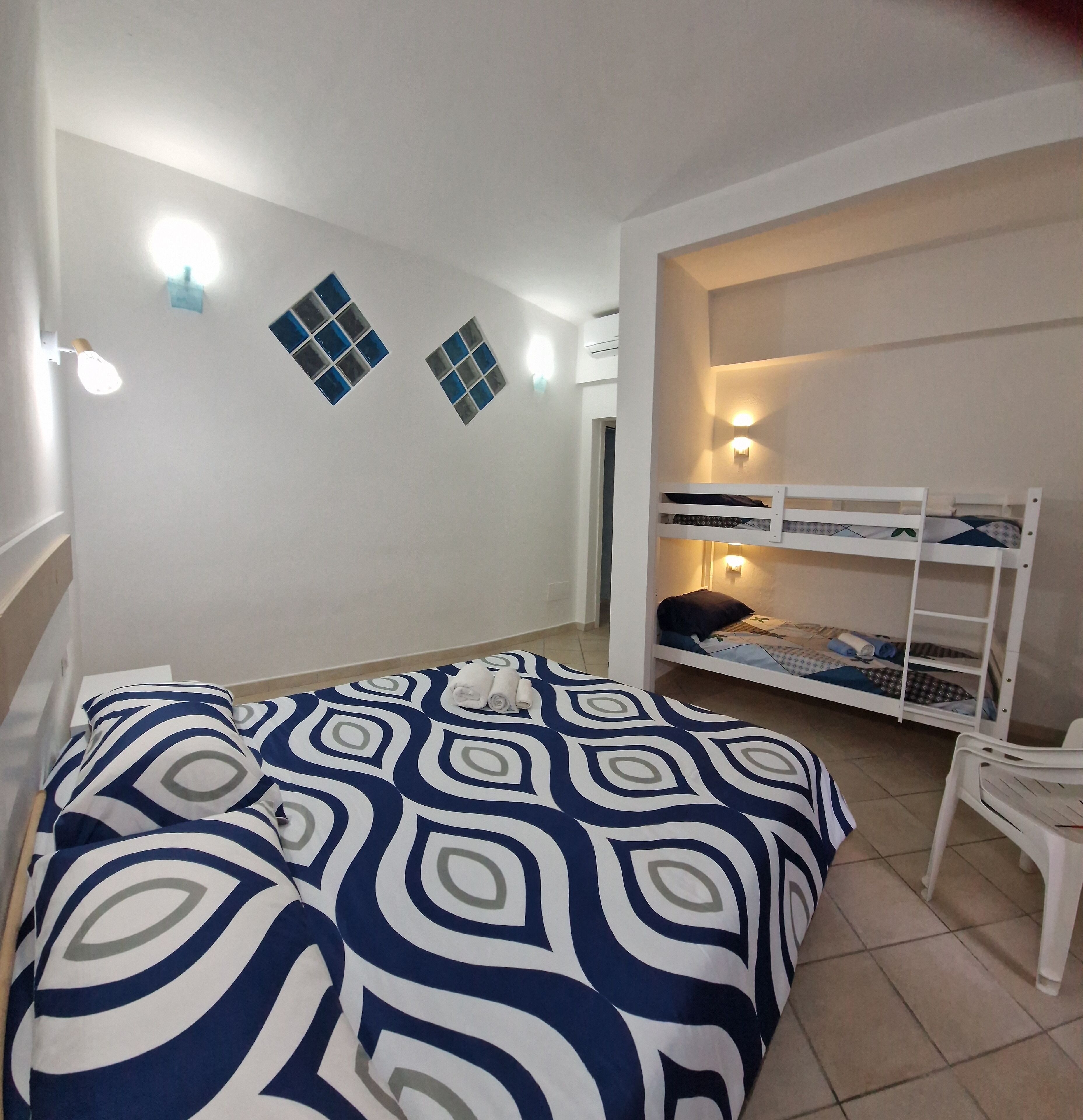 A room with a bed, a bunk bed, a chair, and wall-mounted lights.