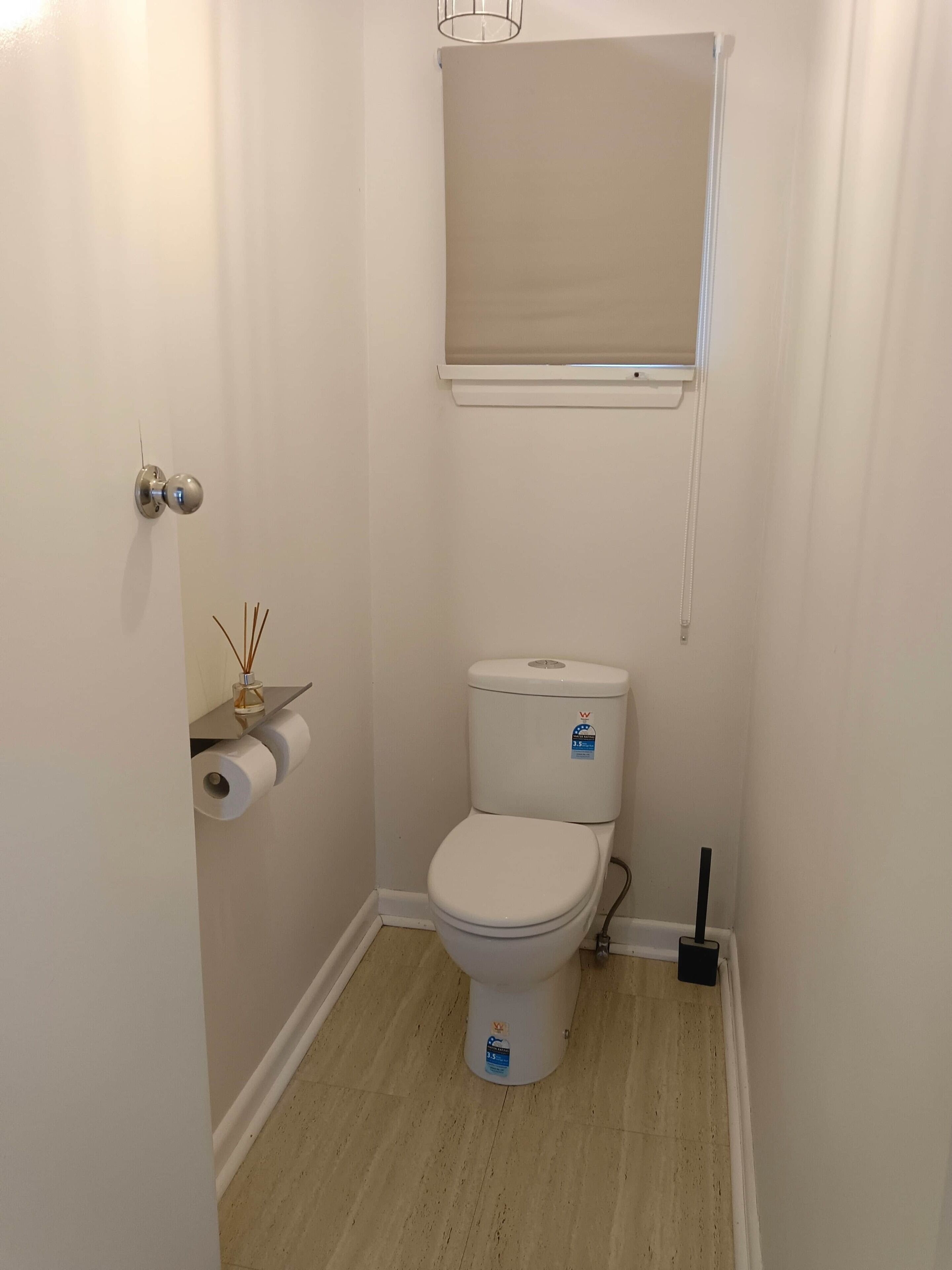 Ein kleines, weißes Badezimmer mit Toilette, Toilettenpapierhalter, Fenster mit Jalousie und Tür mit rundem Griff.