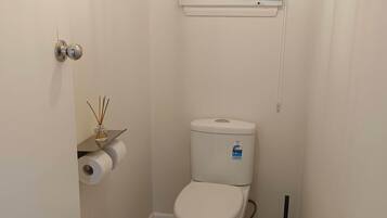 Ein kleines, weißes Badezimmer mit Toilette, Toilettenpapierhalter, Fenster mit Jalousie und Tür mit rundem Griff.
