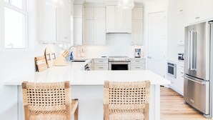A modern kitchen with white cabinets, stainless steel appliances, and a large island with woven chairs. - New Oceanfront Home | Private Beach Access (Topsail Beach)