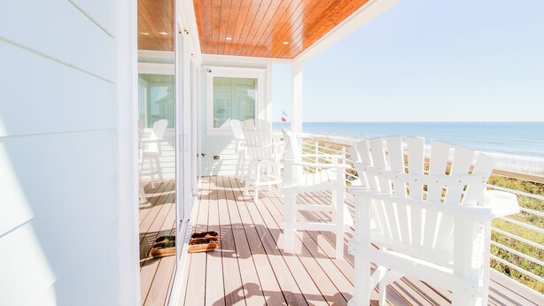 A wooden deck with white chairs and a glass door leading to a beach view. - New Oceanfront Home | Private Beach Access (Topsail Beach)