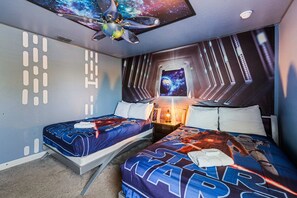 Une chambre avec deux lits, un ventilateur de plafond, un téléviseur fixé au mur et une fenêtre donnant sur l’extérieur.