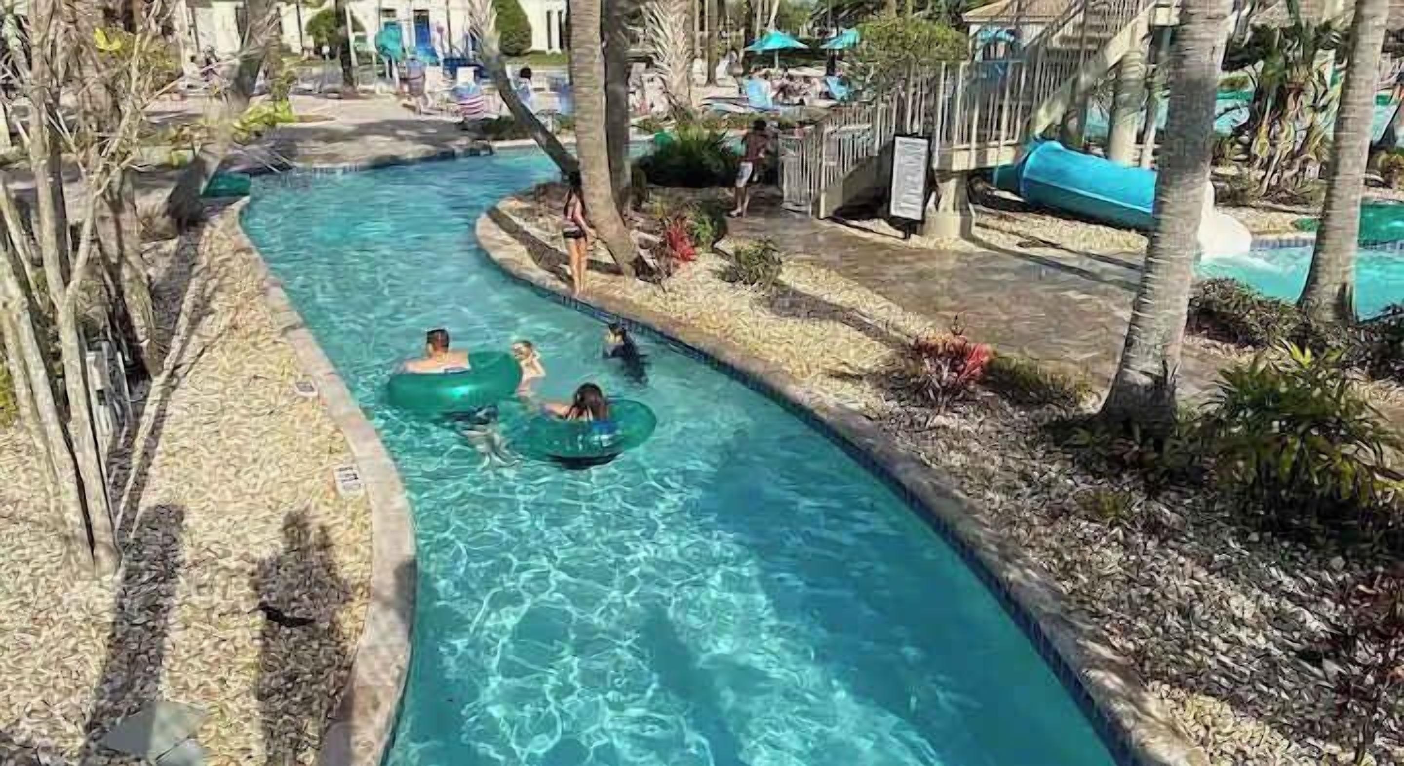 A swimming pool with people on inner tubes, surrounded by palm trees and lounge chairs.