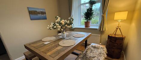 Un comedor con una mesa de madera, individuales, un florero con flores, un banco, una lámpara y una ventana con cortinas.