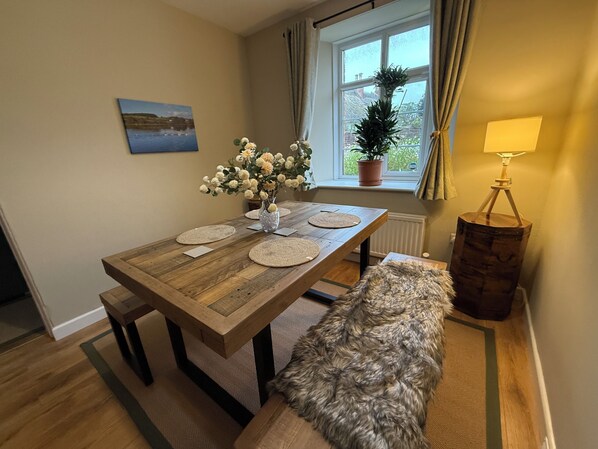 A dining area with a wooden table, placemats, a vase with flowers, a bench, a lamp, and a window with curtains.