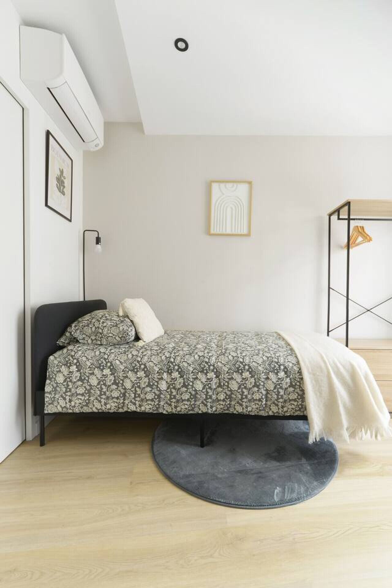 Une chambre à coucher moderne avec un lit, un tapis rond, une table de chevet, une lampe et un porte-manteau.