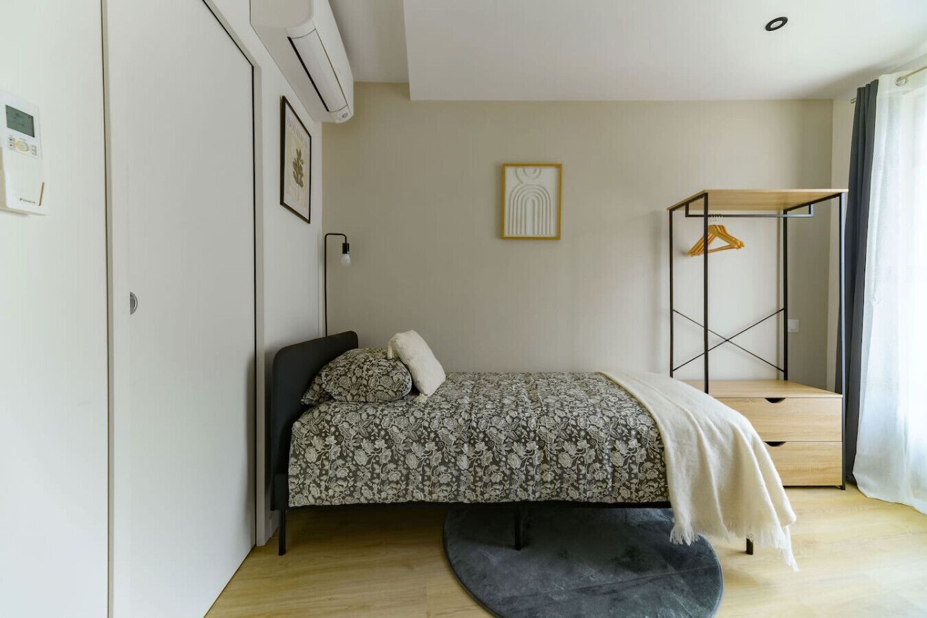 A bedroom with a bed, a bedside table, a hanging light, a wall-mounted air conditioner, and a window with curtains.