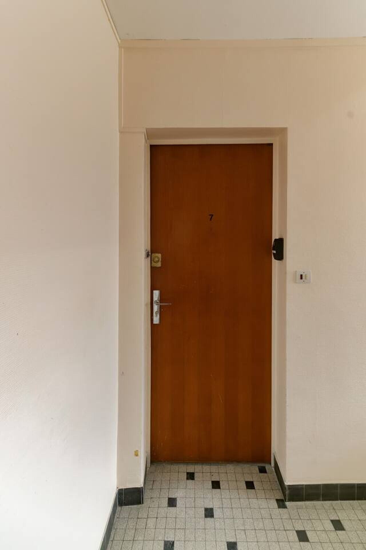 Une porte en bois fermée, dotée d’un clavier numérique pour l’ouverture, d’un interphone fixé au mur et d’un sol carrelé.