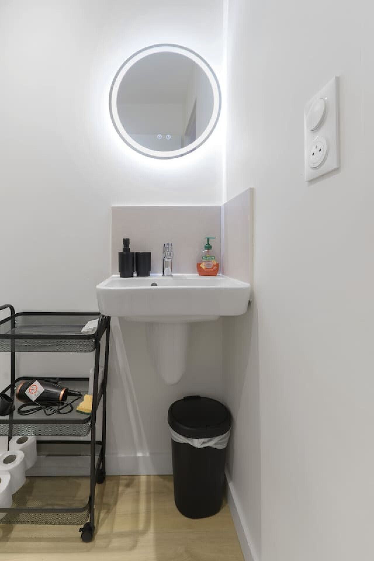 Une petite salle de bain avec un lavabo blanc, un miroir, une poubelle noire et une étagère en métal où sont rangés les articles de toilette.