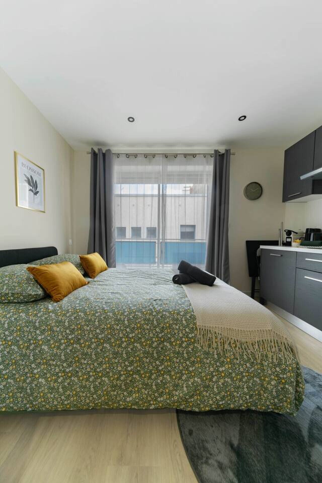 A bedroom with a bed, a small desk, a window with curtains, and a view of buildings outside.
