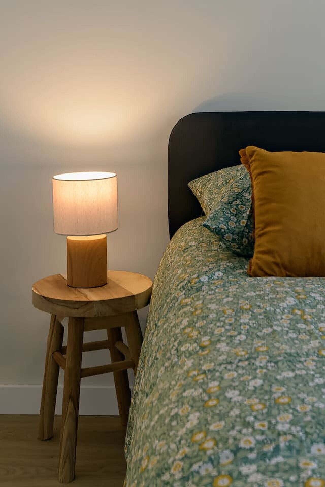 A bed with floral bedding and a mustard-colored pillow, a wooden nightstand with a lamp, and a dark headboard.