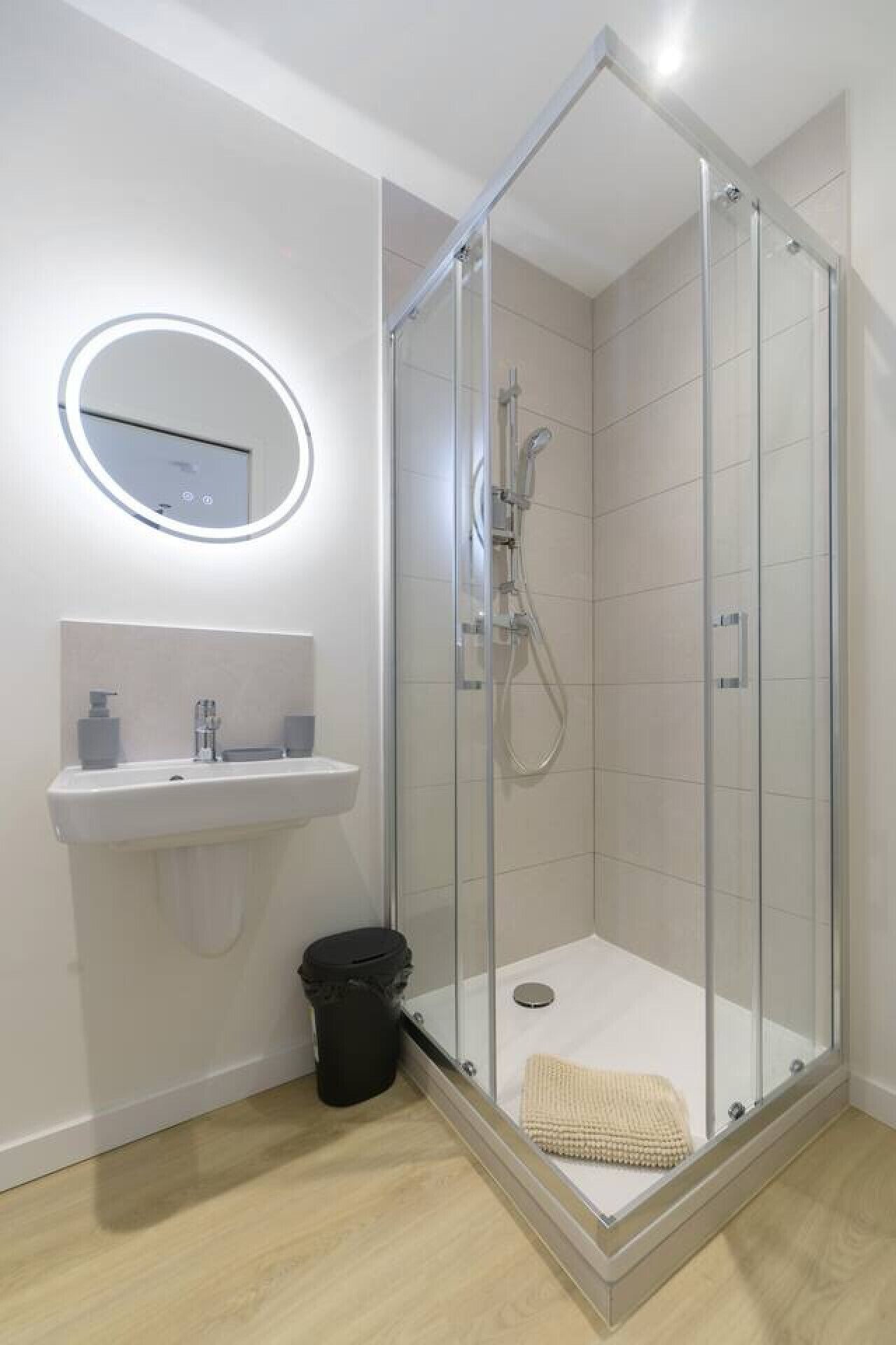 A modern bathroom with a glass-enclosed shower, a round mirror, a sink, and a trash bin.