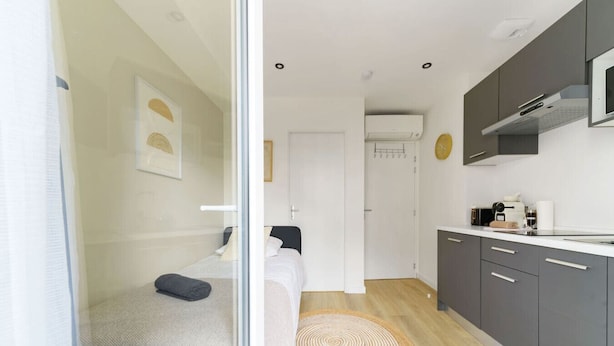 A modern, compact room with a bed, a small kitchenette, and a door leading to another room.
