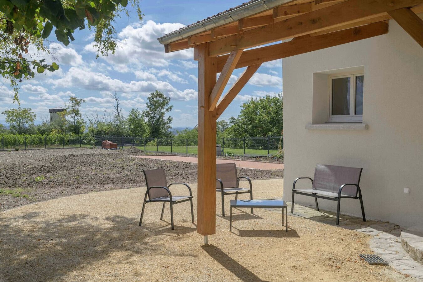 A covered outdoor seating area with four metal chairs and a table, a white building with a window, and a view of a landscaped garden and distant trees.