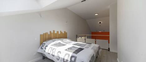 Un dormitorio con techo inclinado, cama con colcha estampada, cabecera de madera y pequeño rincón de escritorio con silla.