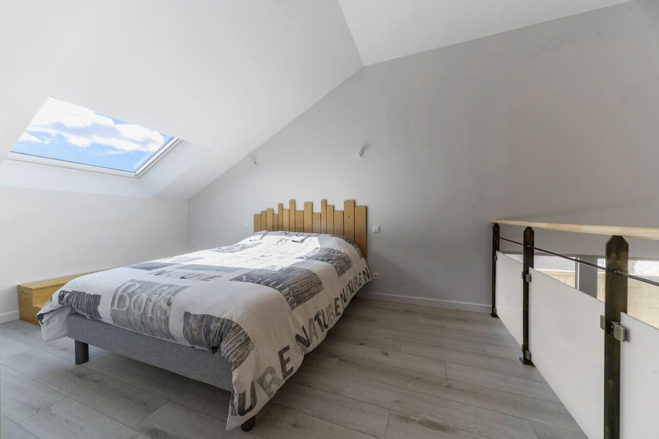Quarto com teto inclinado, cama com colcha estampada, cabeceira de madeira e claraboia.