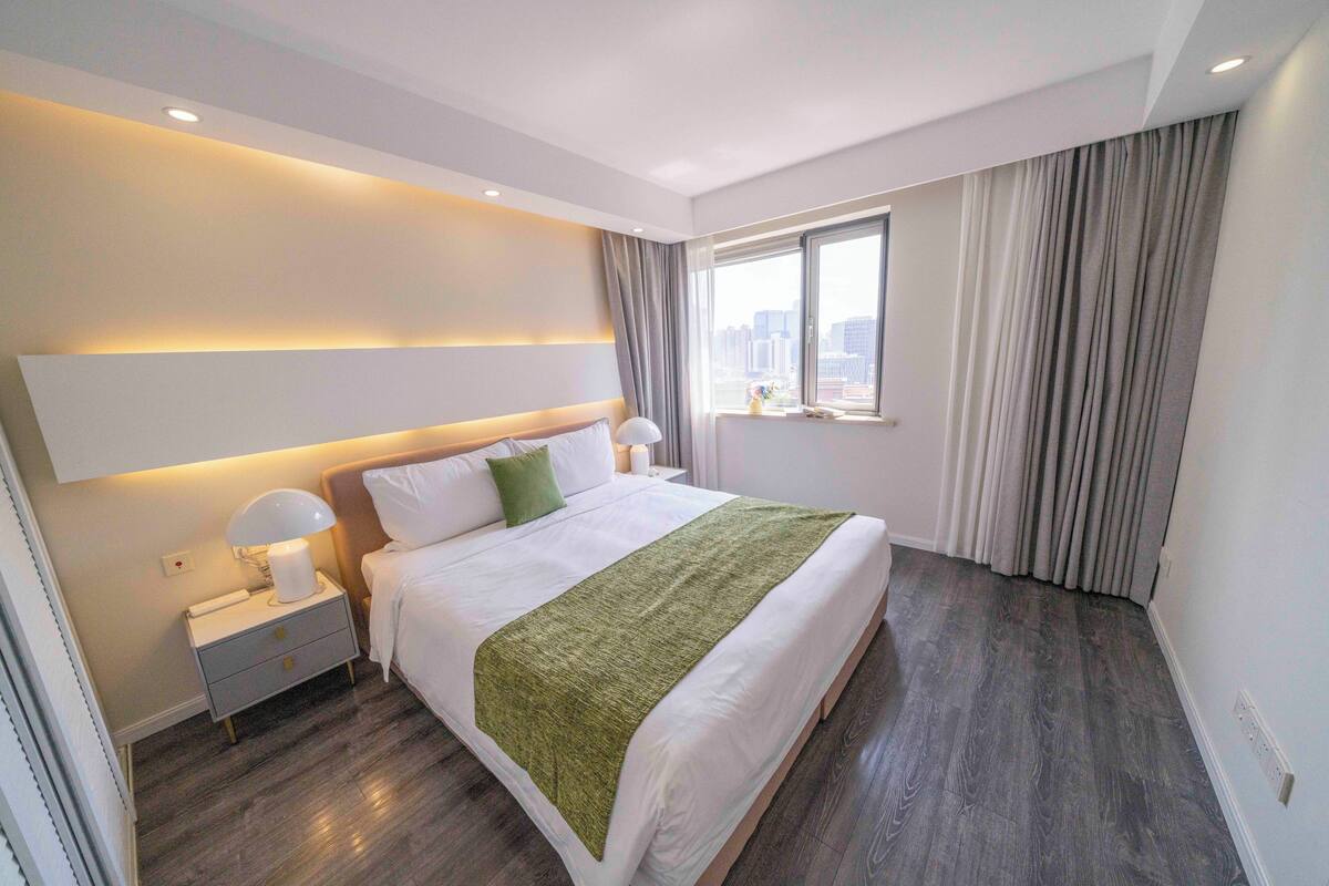 Ein modernes Hotelzimmer mit einem großen Bett, Nachttischlampen, einem Beistelltisch, einem Fenster mit Vorhängen und Blick auf die Stadt.