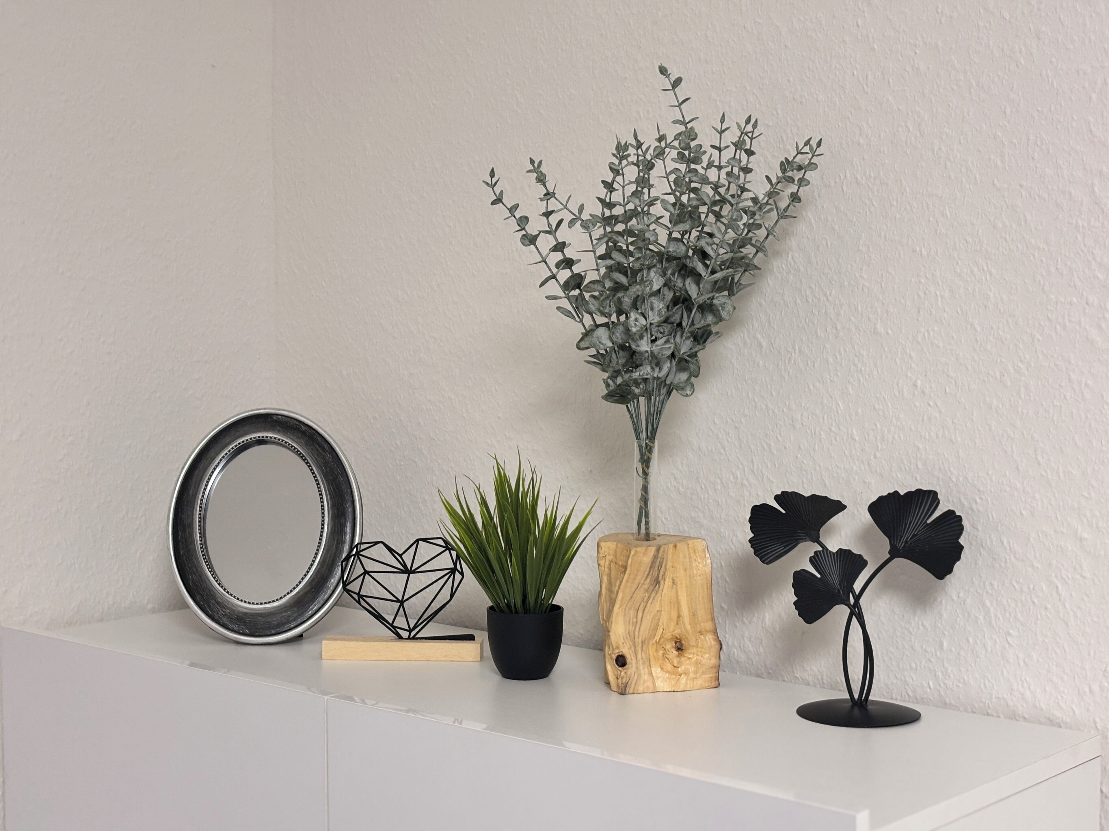Een witte consoles met een spiegel, een geometrisch zwart sculptuur, een potplant, een houten vaas en een zwart decoratief object.
