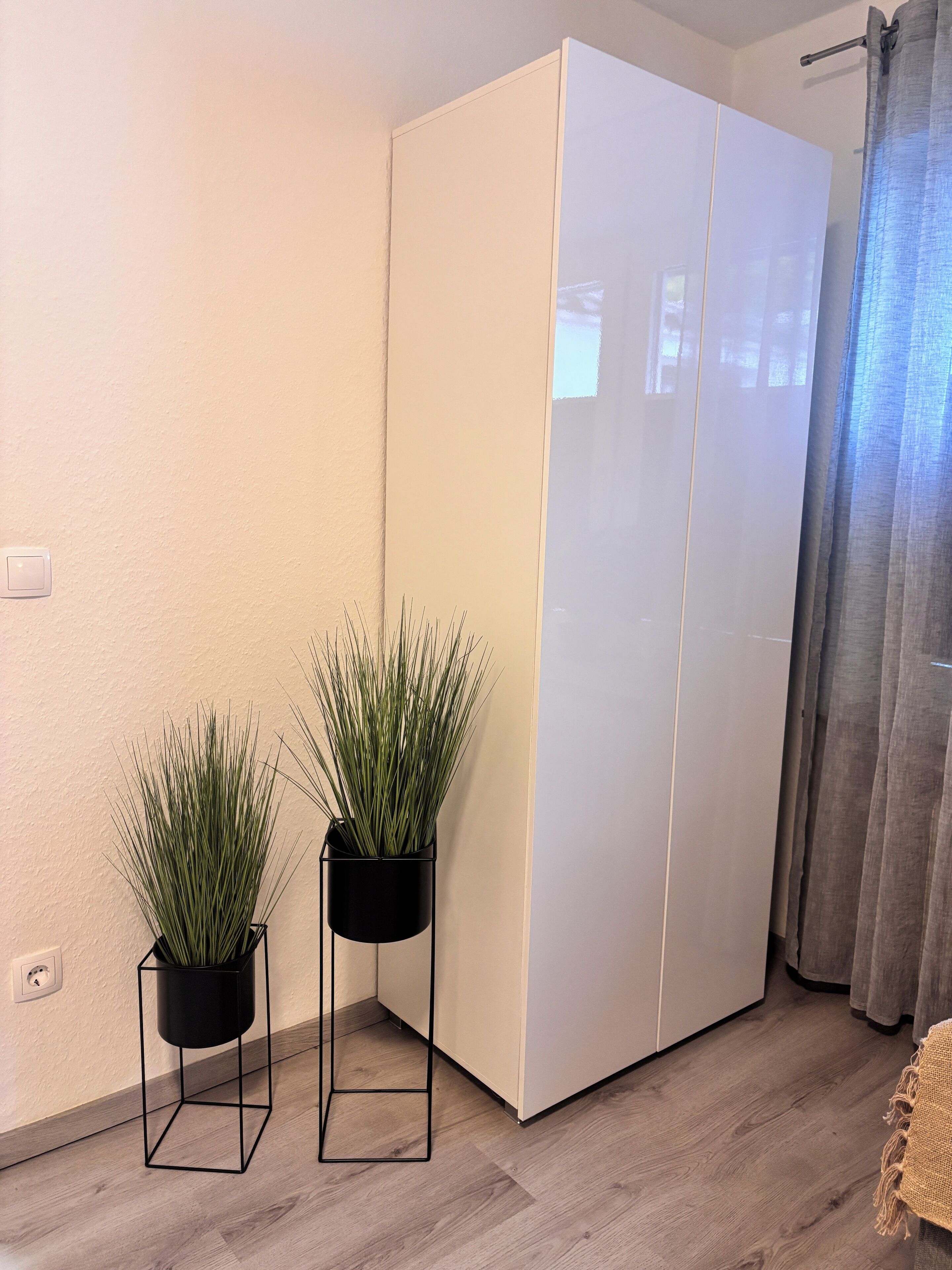 Una habitación con un armario blanco, dos macetas cuadradas negras con plantas verdes, suelo de madera y una cortina gris.