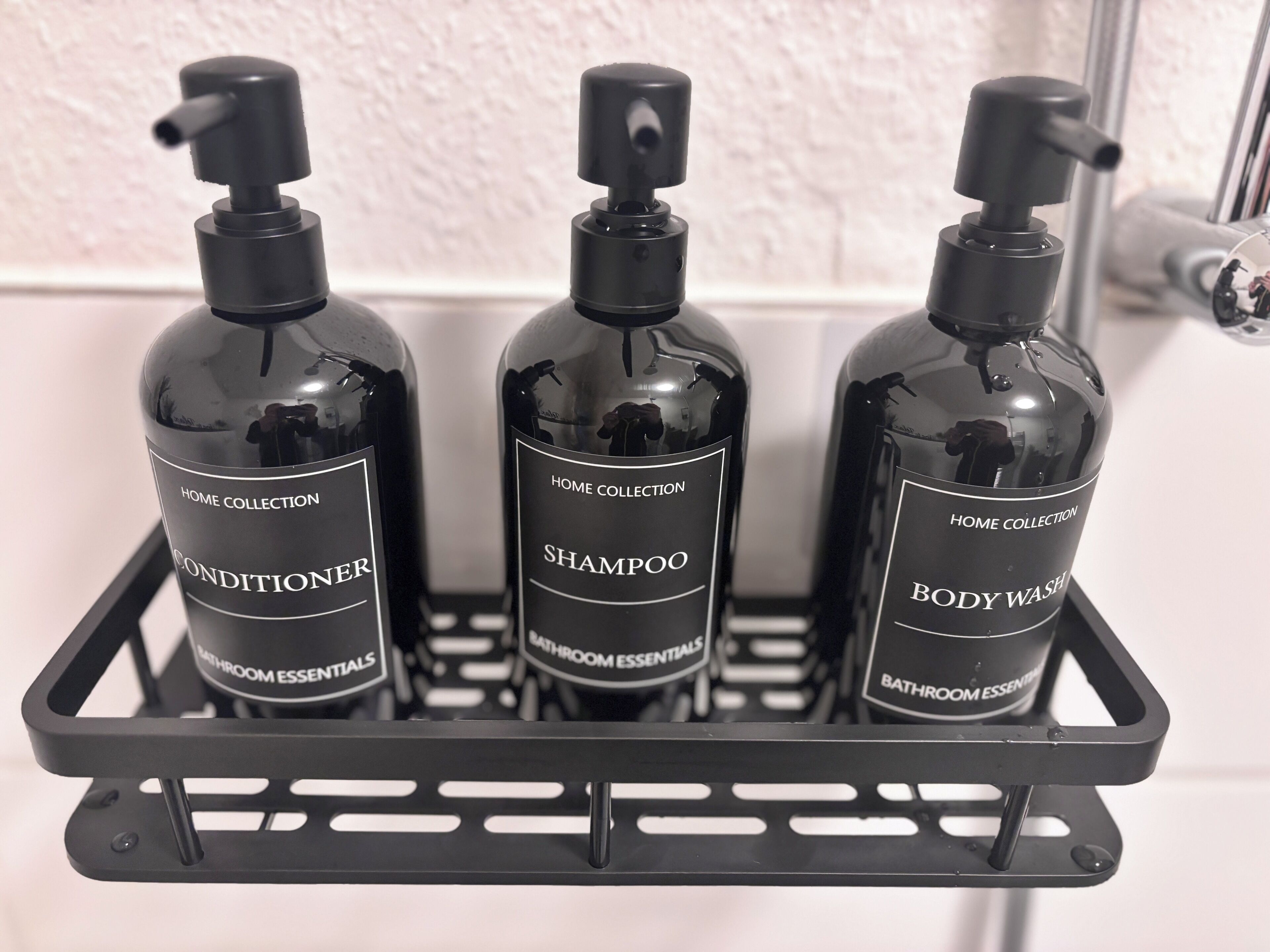 Three black bottles with pump dispensers on a black rack. Each bottle is labeled with a different bathroom essential: conditioner, shampoo, and body wash.