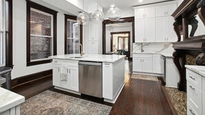 A modern kitchen with white cabinetry, a central island with a dishwasher, and a large window with a brick wall. - Historic-11BR/7.5BA-Parking+By Metro+Chefs Kitchen (Washington)