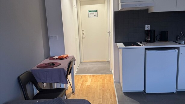 A compact room with a bed, a small dining table with a chair, a kitchenette, and a door leading to another room. - Apartment 17 in Sollentuna (Sollentuna)