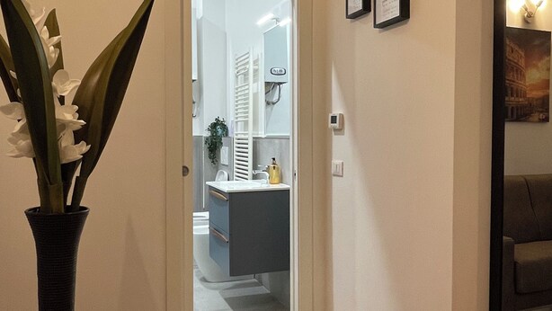 A hallway with a door leading to a bathroom, a framed picture on the wall, and a vase with flowers. - Enjoy Rhome, your apartment just steps from the Basilica of Santa M. Maggiore (Roma)