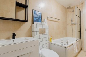 Ein Badezimmer mit weißem Waschbecken, Toilette und Badewanne, einem Handtuchhalter, einer Dusche mit Glastür und einem gerahmten Bild an der Wand.