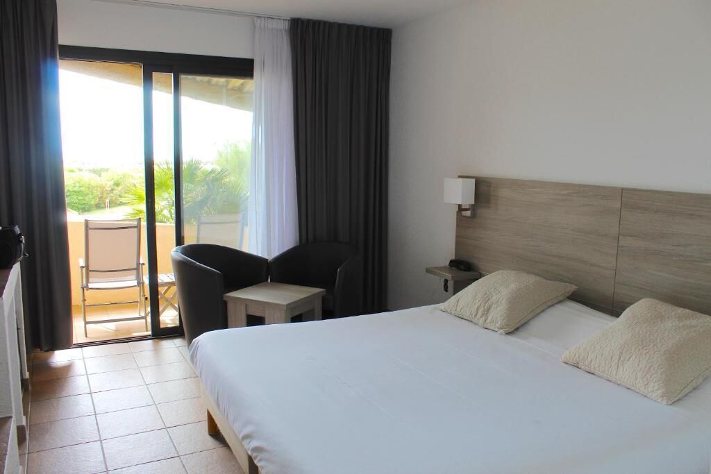 A hotel room with a bed, a chair, a small table, a nightstand, and a view of the outdoors through a sliding door.