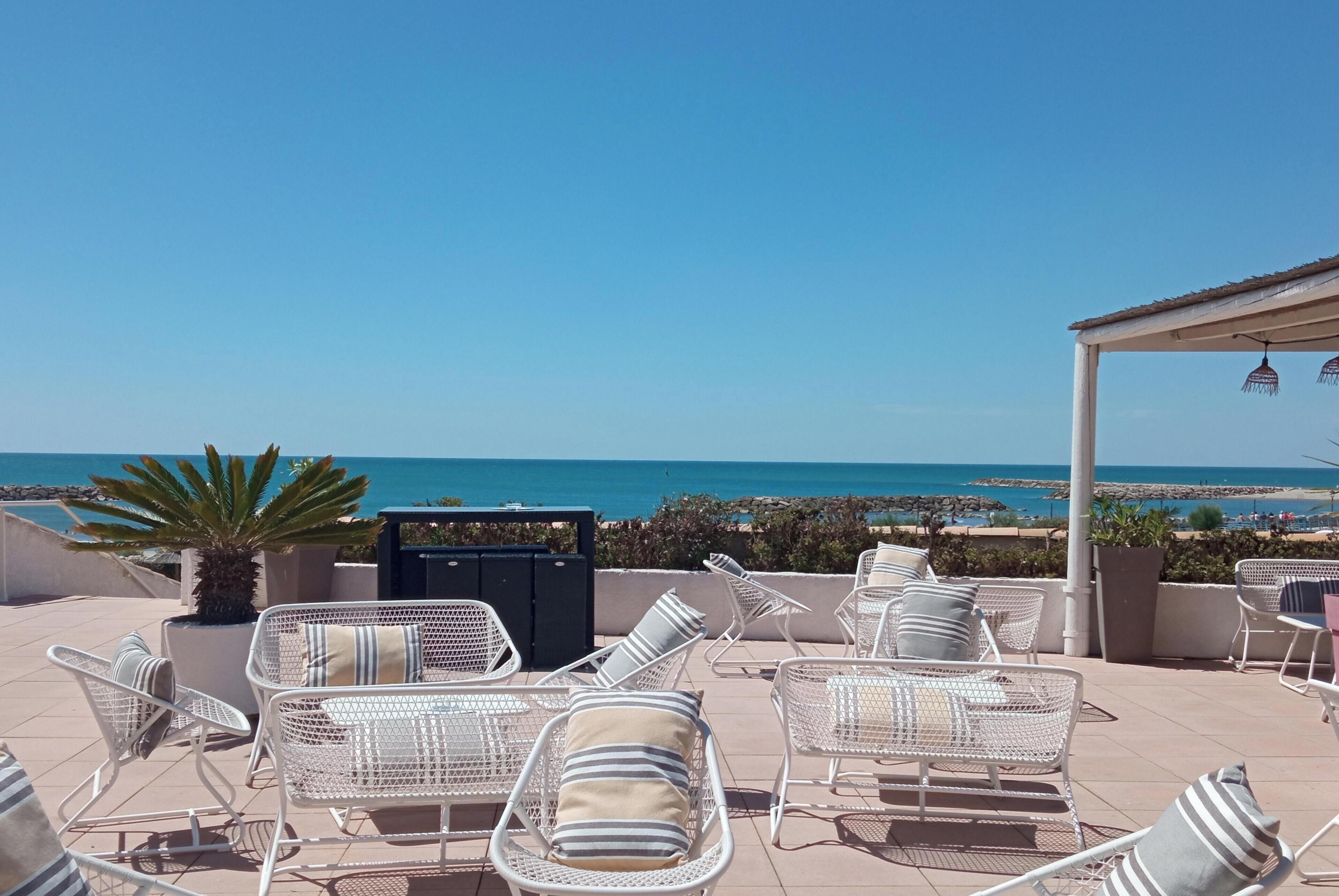 Una terraza con sillas y una mesa de metal blanco, con vista a una playa y el mar.
