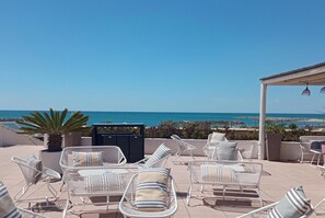 A terrace with white wicker chairs and a table, overlooking a beach with clear blue water. - Thalacap Camargue (Saintes-Maries-de-la-Mer)