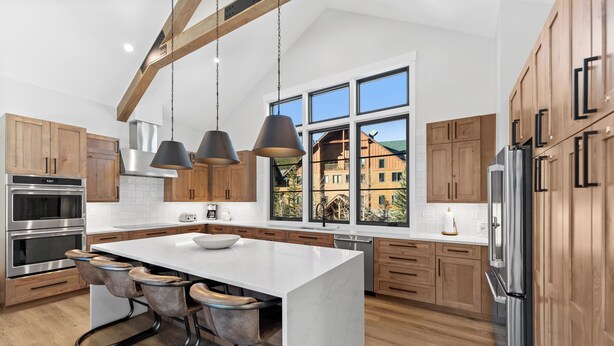 A modern kitchen with a large island, stainless steel appliances, wooden cabinets, and large windows offering a view of a mountain cabin. - Luxury Mountain Retreat - Private Hot Tub and Walk to Lifts (Keystone)