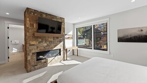 A modern bedroom with a stone fireplace, a bed, a desk, and a large window with a view of a cabin. - Luxury Mountain Retreat - Private Hot Tub and Walk to Lifts (Keystone)