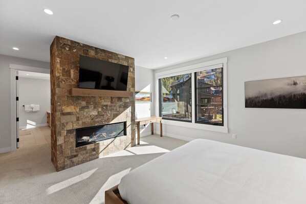 A modern bedroom with a stone fireplace, a bed, a desk, and a large window with a view of a cabin. - Luxury Mountain Retreat - Private Hot Tub and Walk to Lifts (Keystone)