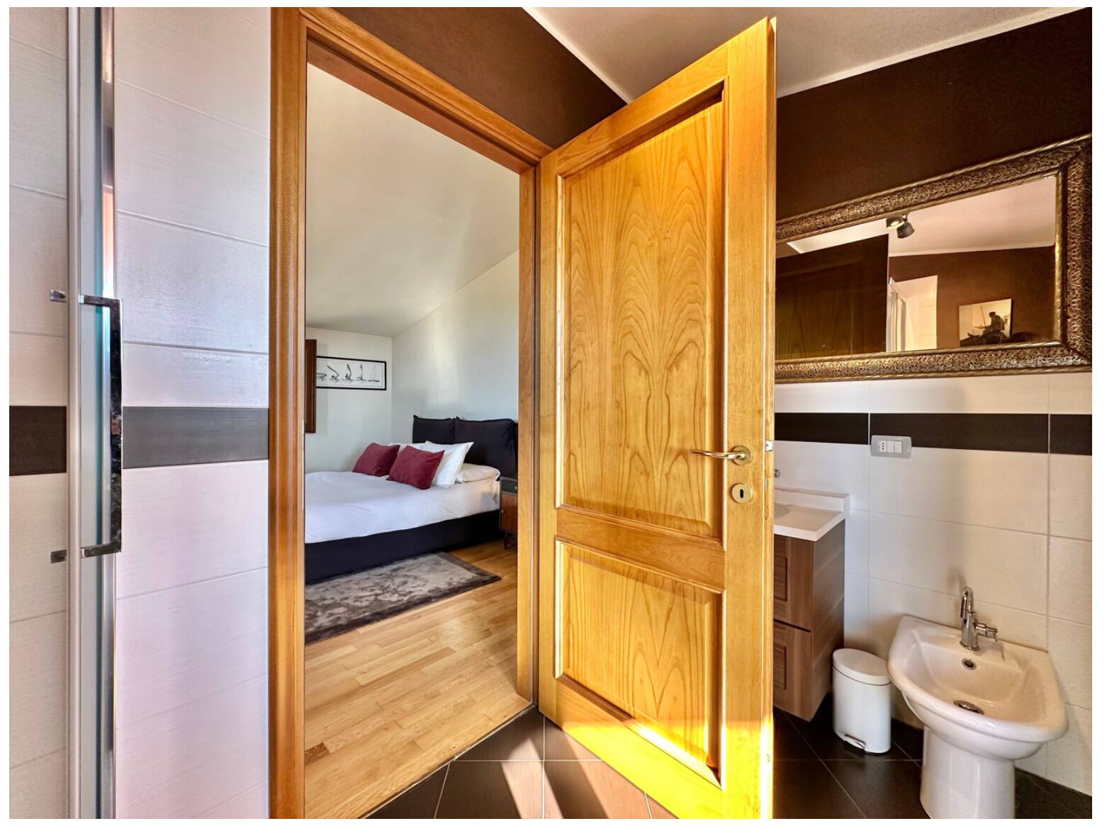 A hotel room with a wooden door, a bed with red and white bedding, a bedside table, a mirror, a sink, and a trash bin.