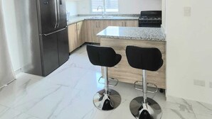 Une cuisine moderne dotée d’un réfrigérateur en acier inoxydable, de meubles en bois, d’un plan de travail en granit et de deux tabourets de bar noirs.