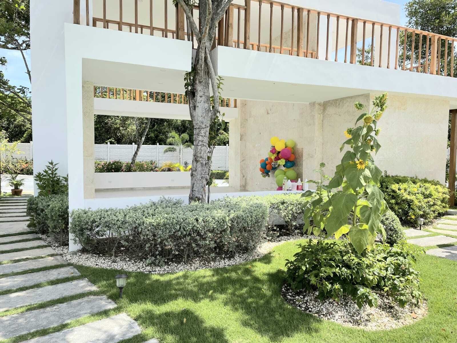 A modern building with a white facade, a wooden balcony, and a garden with various plants and a colorful balloon decoration.