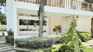 Un bâtiment moderne à la façade blanche, doté d’un balcon en bois et d’un jardin où poussent différentes plantes, agrémenté d’une décoration festive composée de ballons de couleurs vives.