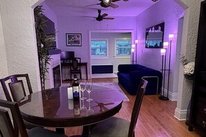 A dining area with a round table, chairs, and a bottle of wine. There is a sofa and a floor lamp in the background. - Charming 2 BR house with pool and hot tub walking distance to the beach/Las Olas (Fort Lauderdale)