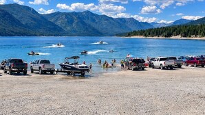 Un lac avec des bateaux, une plage où sont garés des véhicules, et des montagnes en arrière-plan.