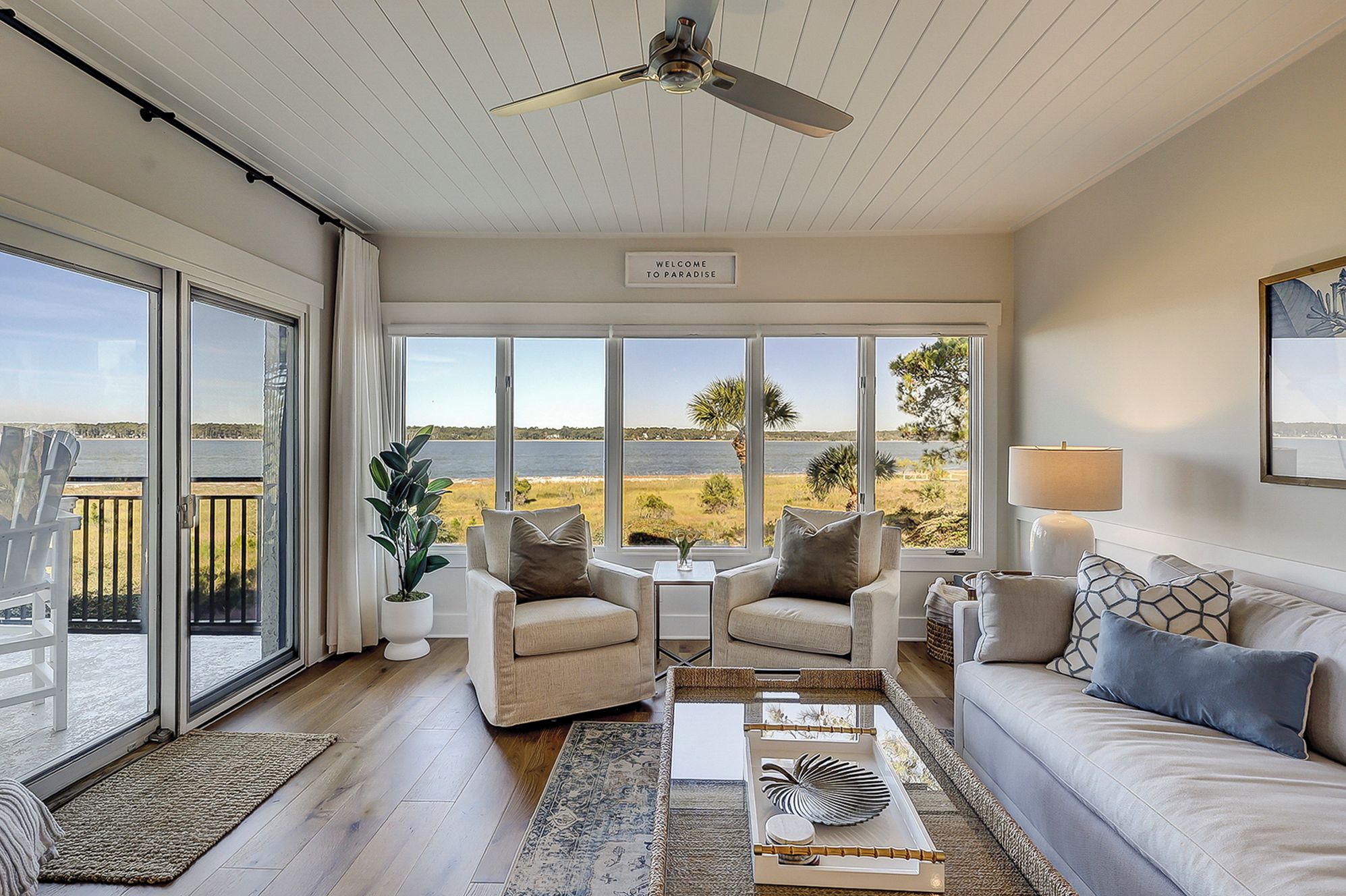 A spacious living room with a view of the sea, a ceiling fan, and a sign that reads "Welcome to Paradise".