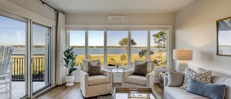 A spacious living room with a view of the sea, a ceiling fan, and a sign that reads "Welcome to Paradise".