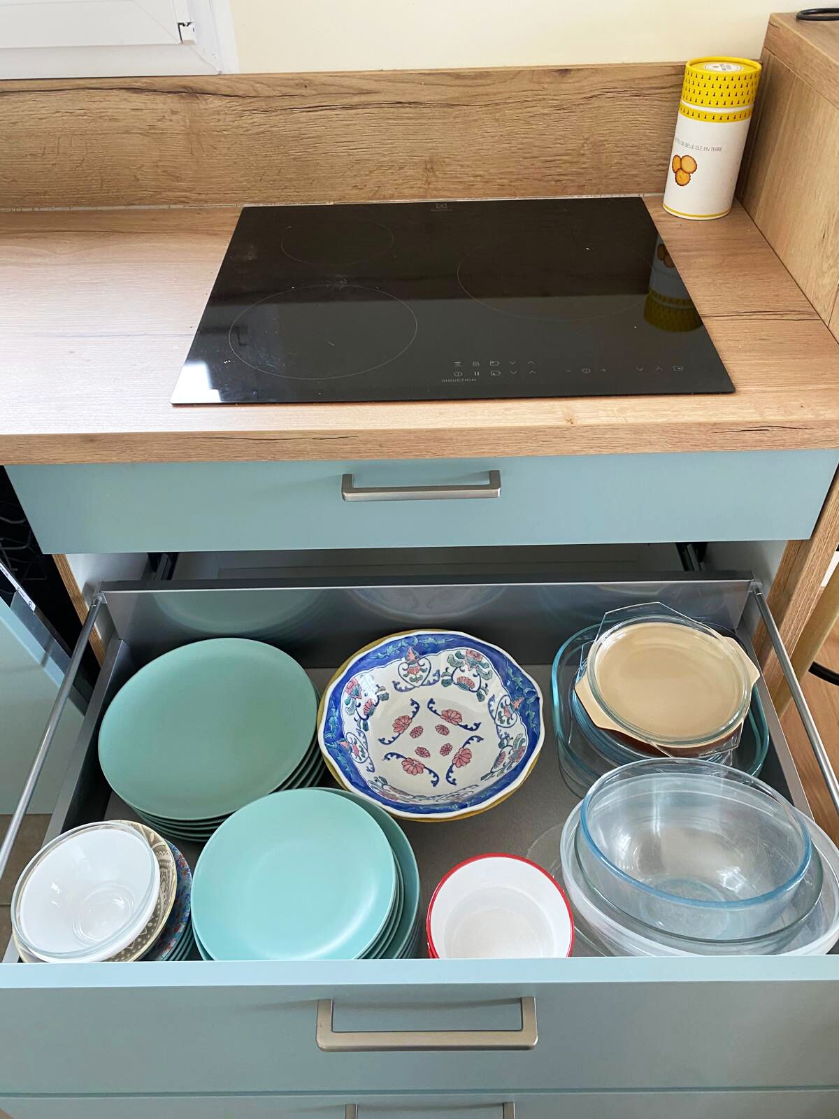 Un tiroir de cuisine surmonté d’une plaque de cuisson à induction noire, contenant divers assiettes et bols.