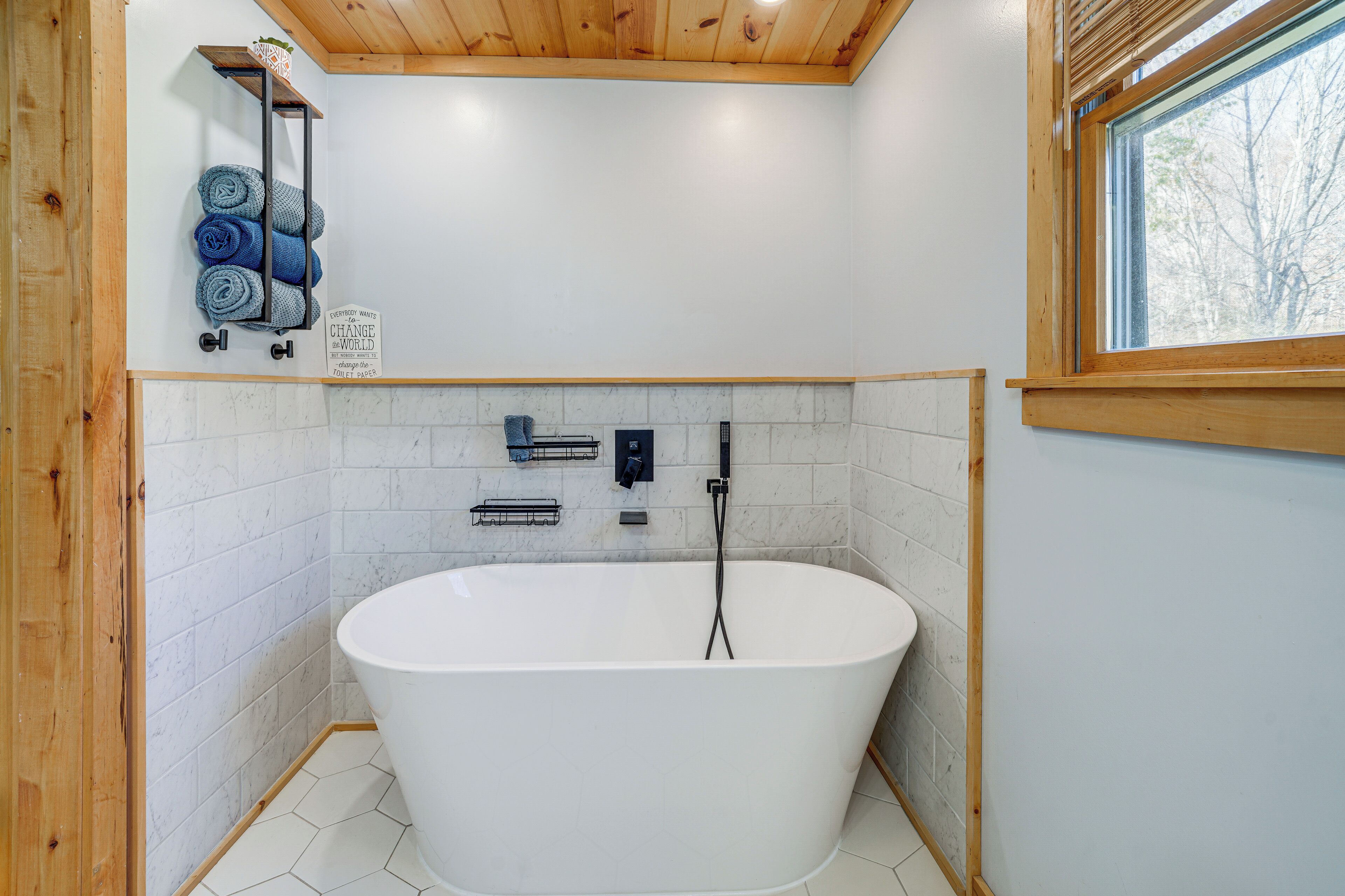 Un baño moderno con una bañera independiente, techo de madera y una ventana que aporta luz natural.