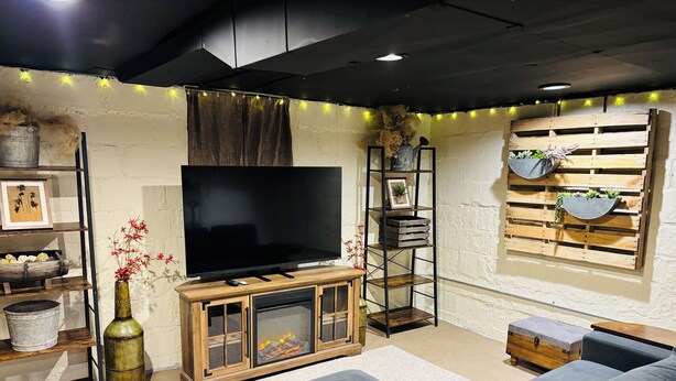 Ein gemütliches Wohnzimmer mit einem Flachbildfernseher, einem Kamin, einem grauen Sofa und einem Holzregal mit Pflanzen.