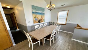 A dining area with a wooden table and white chairs, a painting of a beach scene, and a chandelier.