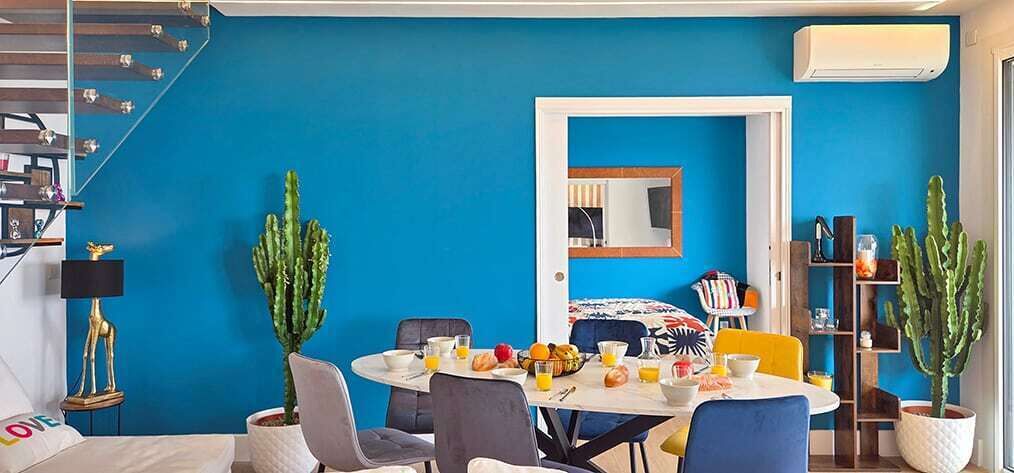 A room with a blue wall, a dining table with chairs, a bed with a colorful headboard, and two potted cacti.