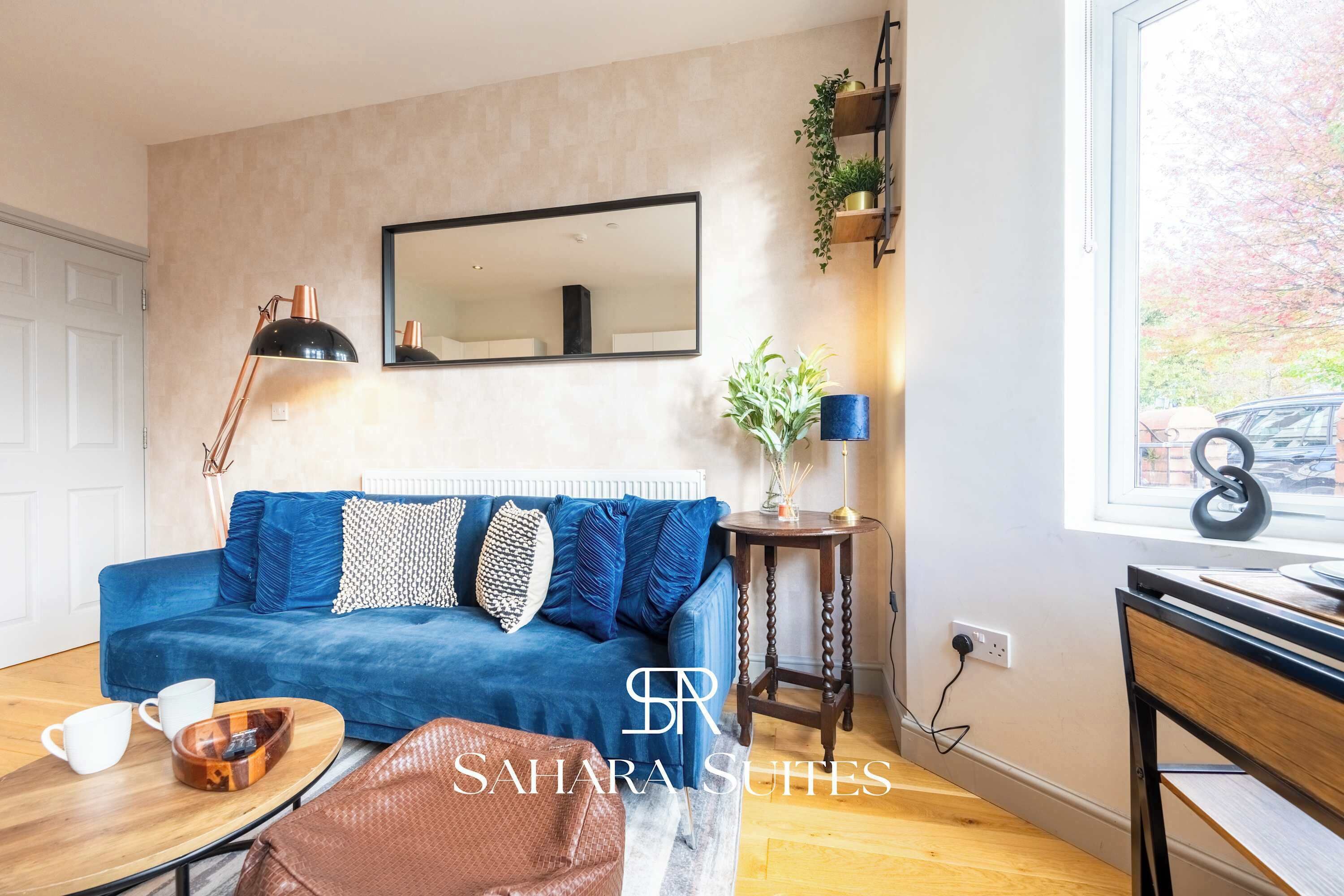 A living room with a blue sofa, a coffee table, a side table with a lamp, a mirror, and a window with a view of trees.