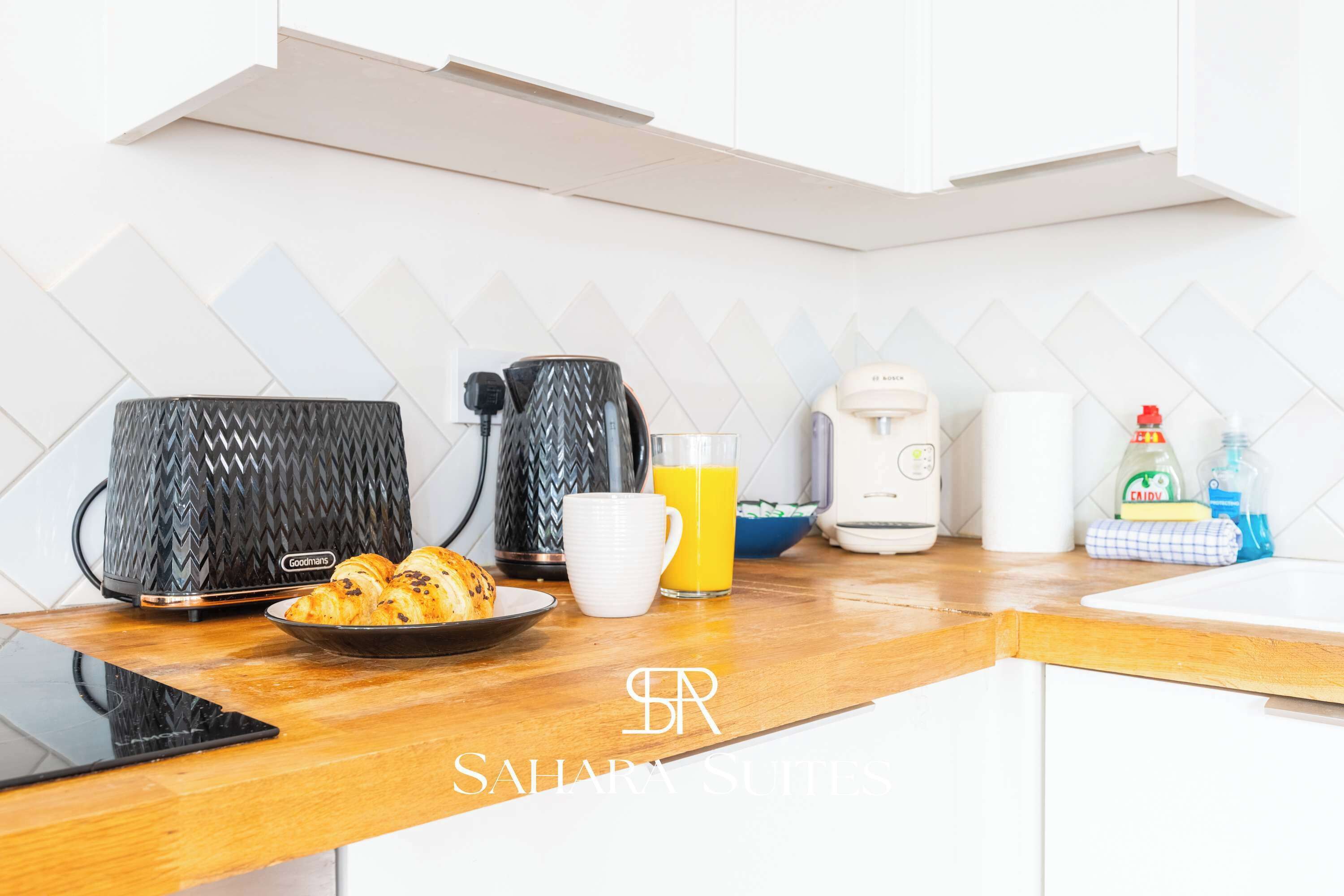 A modern kitchen with a coffee maker, a kettle, a toaster, a plate of croissants, a glass of orange juice, and a tablet on the counter.