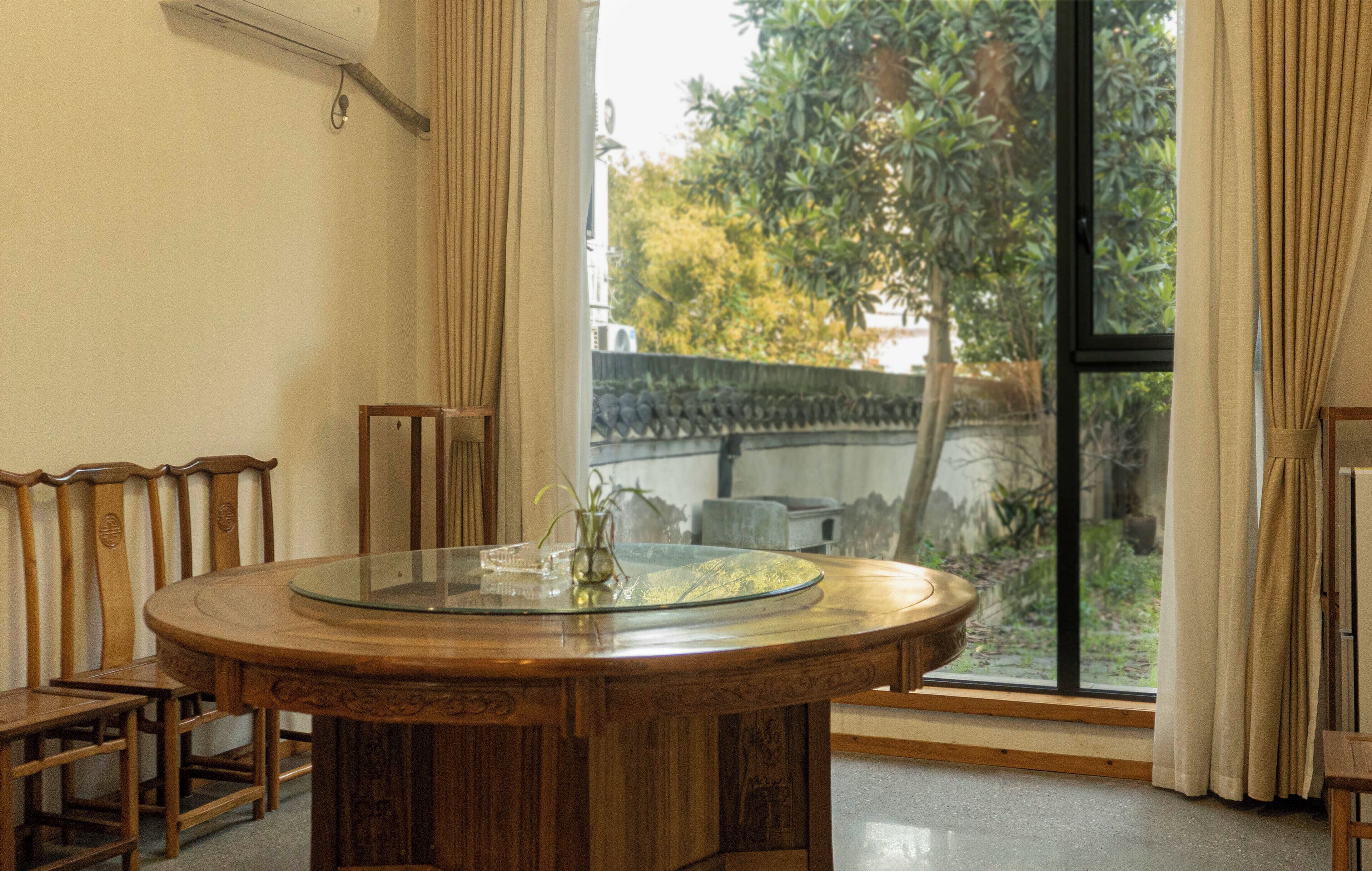 A round wooden dining table with chairs, a glass top, and a vase with a plant.